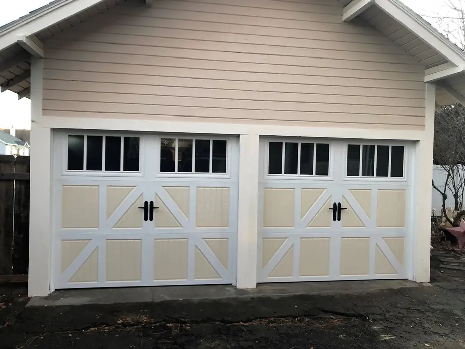 A new pair of almond carriage-house garage doors with window tops and black handles on a tan garage